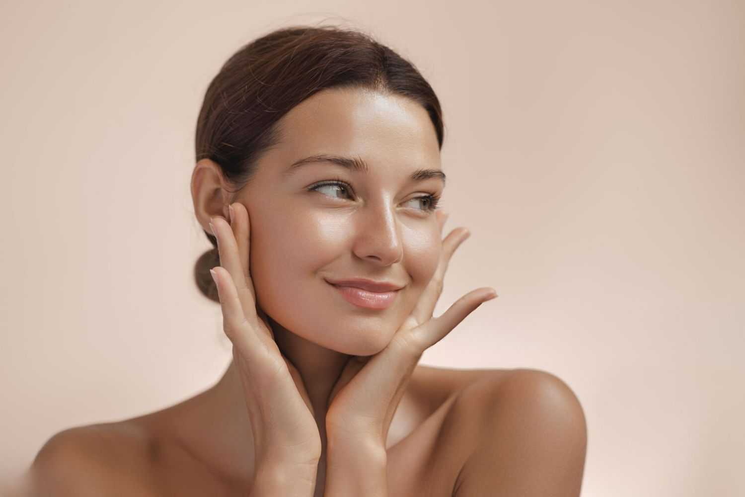 Young woman with a serene expression, hands gently touching her face against a soft background.