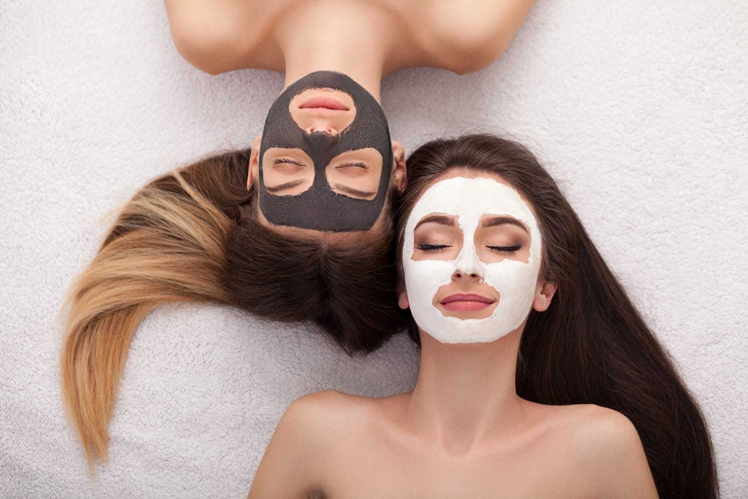 Two women with clay masks relaxing, one with a black mask, the other with a white mask on their faces.