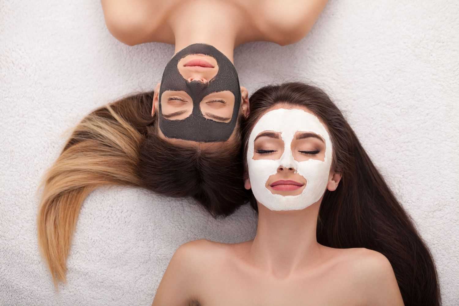 Two women with clay masks relaxing, one with a black mask, the other with a white mask on their faces.
